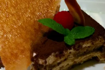 U trozo de tiramisu decorado con cacao, menta y frambuesa, acompañado con barquillo.