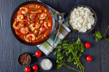 Gumbo de gambas y salchicha con un cuenco de arroz blanco y condimentos sobre pizarra.