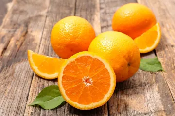 Una naranja cortada, dos naranjas enteras y hojas sobre madera.