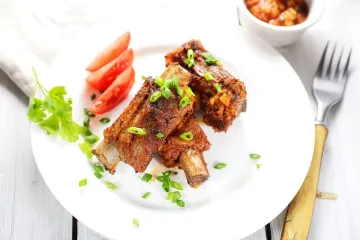 Un plato con costillas de cerdo aromatizadas y fritas acompañadas de tomate y decoradas con cebollino.