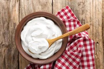 Un cuenco de madera con yogur natural con una cuchara de palo sobre una servilleta de cuadros en una mesa de madera.