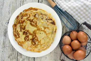 Un palto con crêpes caseras con un batidor de globo y un cuenco con huevos frescos al lado.