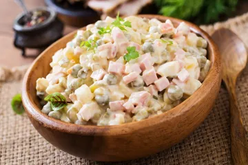 Un cuenco de madera con ensalada Olivier, o ensalada rusa, con patata, zanahoria, guisantes, jamón y pepinillos troceado y mezclados con mayonesa. El cuenco está colocado sobre una superficie de arpillera y hay una cuchara al lado.