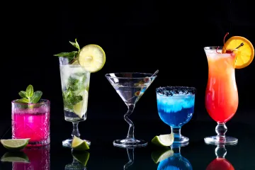 Una fila de cócteles de colores brillantes sobre una mesa de cristal, contra un fondo negro.