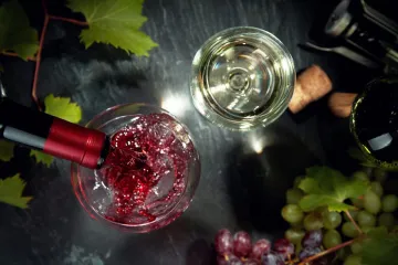 Sirviendo vino tinto en una copa, mientras hay otra copa llena con vino blanco al lado y uvas alrededor, naturaleza muerta.