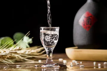 Un chorro de licor blanco chino, baijiu, llenando una copa transparente, con una botella de cerámica detrás que supuestamente también contiene este tipo de licor. 