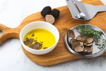 Aceite de oliva en un tazón blanco con rodajas de trufa negra y tomillo en una tabla de cortar, junto a un rallador de trufas.
