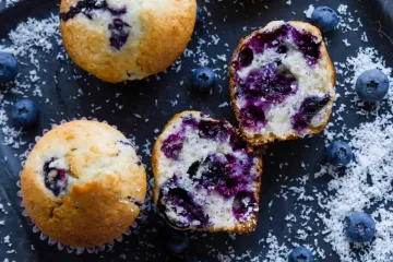 Muffins de arándanos azules sobre una superficie oscura, uno de ellos partido por la mitad mostrando su interior esponjoso con abundantes arándanos, rodeados de frutos frescos y coco rallado.
