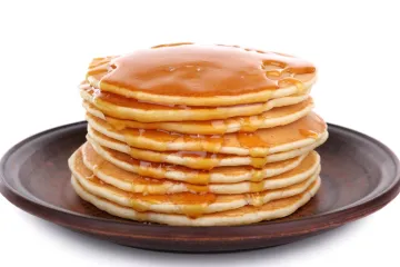 Torre de tortitas americanas doradas cubiertas con sirope de arce, servidas en un plato de cerámica oscura sobre un fondo blanco.