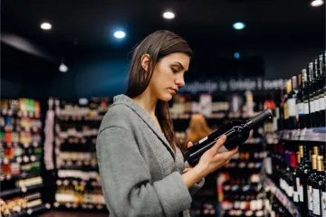 Mujer joven leyendo la etiqueta de una botella de vino tinto en el pasillo de una tienda de vinos.