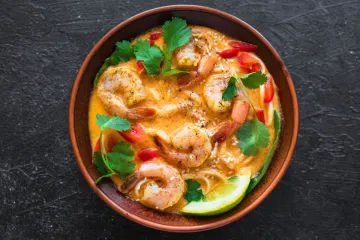 Cuenco de barro con laksa de gambas en un caldo cremoso anaranjado, con fideos, tiras de pimiento rojo, hojas de cilantro, semillas de sésamo y una rodaja de lima, visto desde arriba sobre una superficie oscura.