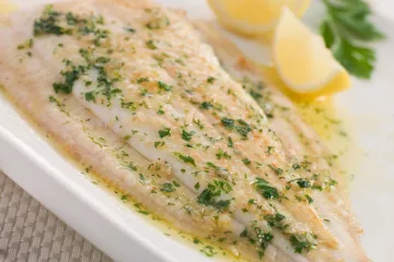 Filete de lenguado a la molinera servido en un plato blanco, napado con mantequilla y perejil picado y acompañado de unos gajos de limón.