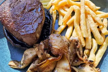 Solomillo de ternera a la plancha sobre un sombrero de portobello, acompañado de setas y alcachofas salteadas y de patatas fritas, en un plato azul.