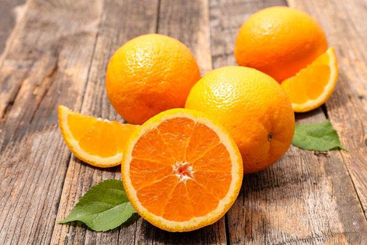 Una naranja cortada, dos naranjas enteras y hojas sobre madera.