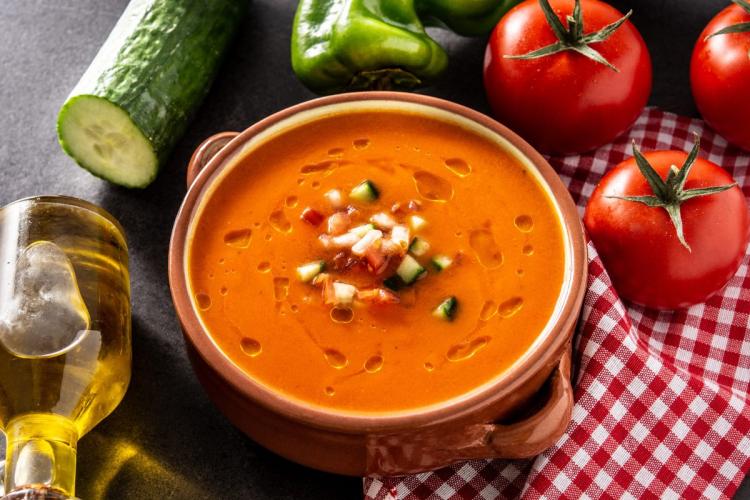 Un cuenco de gazpacho con sus guarniciones típicas rodeado de los ingredientes que se usan para hacerlo.