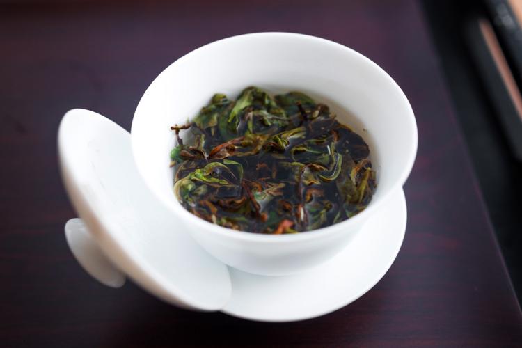 Gaiwan de porcelana blanca con hojas de té en remojo en agua caliente y tapa apoyada a un lado.
