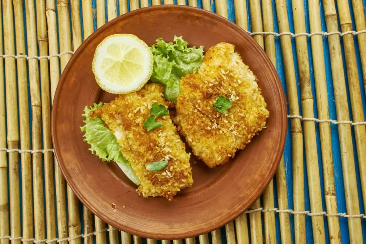 Dos piezas d pescado rebozado en pan y coco rallados y fritas en un plato rústico, acompañado de limón y lechuga.