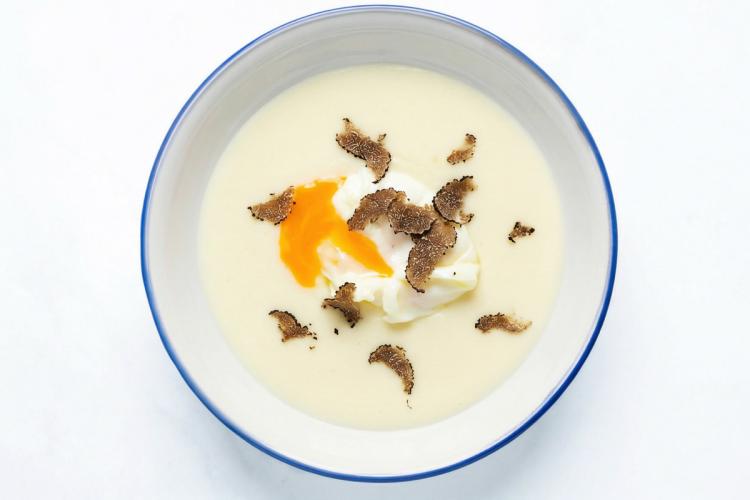 Huevo trufado y escalfado sobre parmentier de patata cremosa en un cuenco blanco de borde azul, visto desde arriba, con yema ligeramente abierta y finas hebras de trufa negra como única decoración.