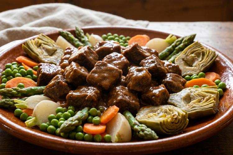 Aguja de ternera a la jardinera con salsa, acompañada de guisantes, zanahorias, nabos, espárragos y alcachofas.