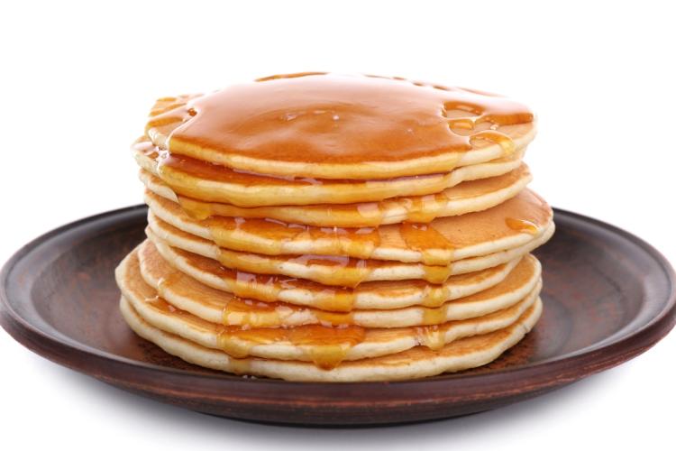 Torre de tortitas americanas doradas cubiertas con sirope de arce, servidas en un plato de cerámica oscura sobre un fondo blanco.