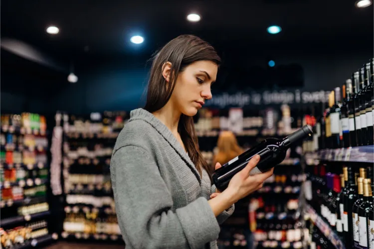 Mujer joven leyendo la etiqueta de una botella de vino tinto en el pasillo de una licorería