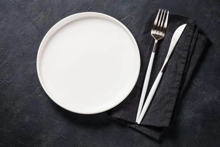 Vista cenital de un plato blanco mate junto a una servilleta de lino negra doblada con un tenedor y un cuchillo de mango blanco encima, sobre una superficie oscura de textura pizarra.
