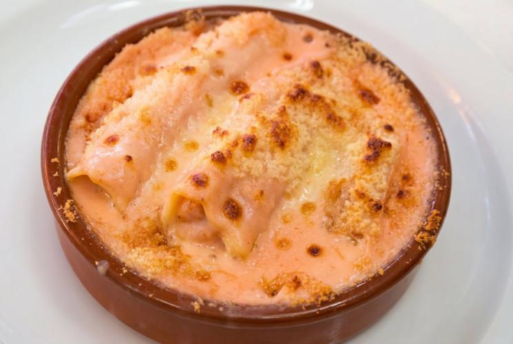 Una porción de canelones de verduras gratinados con bechamel de tomate en una cazuela de barro sobre un plato blanco.
