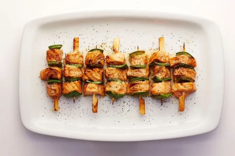 Siete brochetas de salmón teriyaki y pimiento verde a la parrilla, servidas en una fuente blanca y espolvoreadas con pimienta.