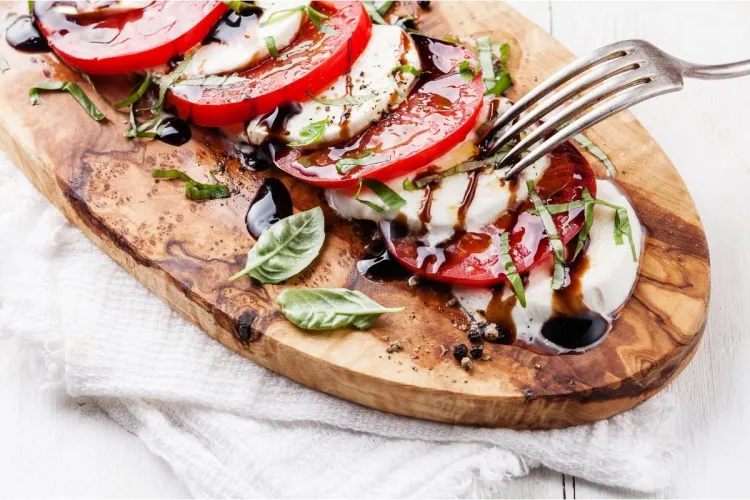 Rodajas de tomate y mozzarella alternadas sobre una tabla de madera, con hojas de albahaca fresca, un chorreado de crema de vinagre balsámico y un tenedor pinchando una de las lonchas.