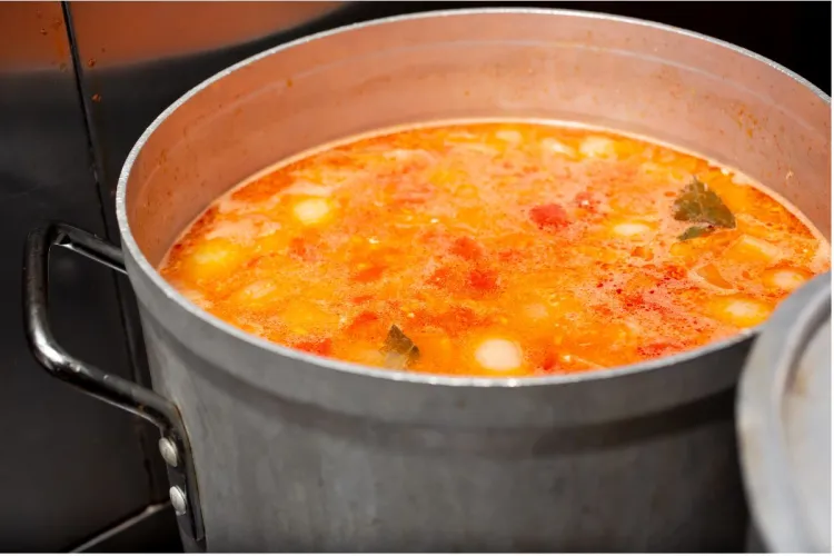 Olla grande de aluminio con una sopa de tomate hirviendo a fuego fuerte, con hojas de laurel y trozos de verdura flotando en el caldo naranja.