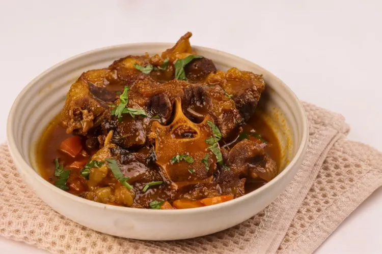 Cuenco de cerámica clara con rabo de toro braseado en una salsa espesa y brillante, con trozos de zanahoria y perejil picado por encima, sobre un paño de cocina beige.