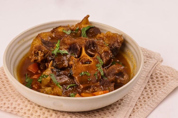 Cuenco de cerámica clara con rabo de toro braseado en una salsa espesa y brillante, con trozos de zanahoria y perejil picado por encima, sobre un paño de cocina beige.