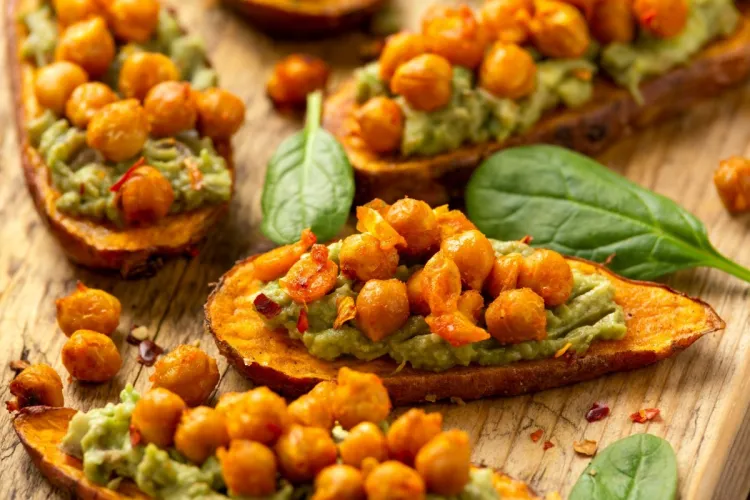 Rodajas de batata asada cubiertas con guacamole y garbanzos tostados y especiados, decoradas con hojas de espinaca baby y copos de guindilla.