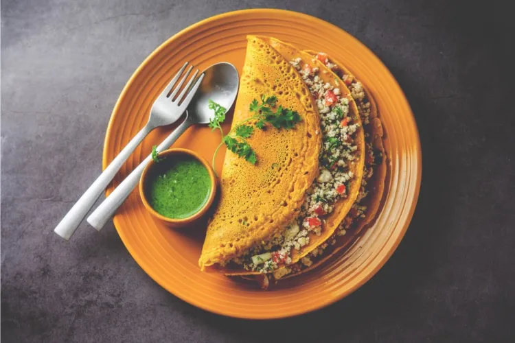 Crepes indios de harina de garbanzos (besan chilla) doblados y rellenos, servidos en un plato naranja con un cuenco de chutney verde, cilantro fresco, tenedor y cuchara.