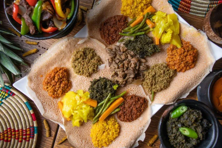 Torta de pan de injera cubierta con montoncitos de guisos, lentejas, verduras y carne especiada, vista desde arriba, sobre una mesa con cestería etíope