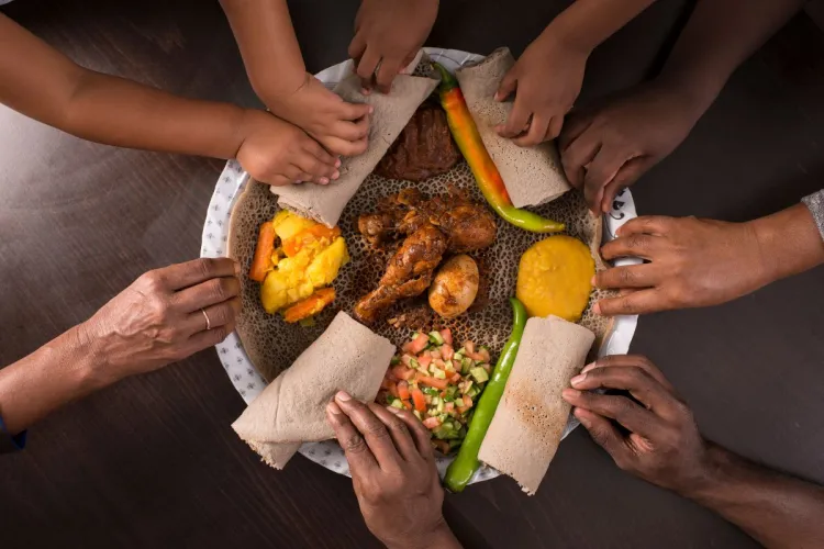 Varias manos comparten una bandeja etíope de injera con pollo, guisos, verduras al curry, huevo y guindillas, vista desde arriba