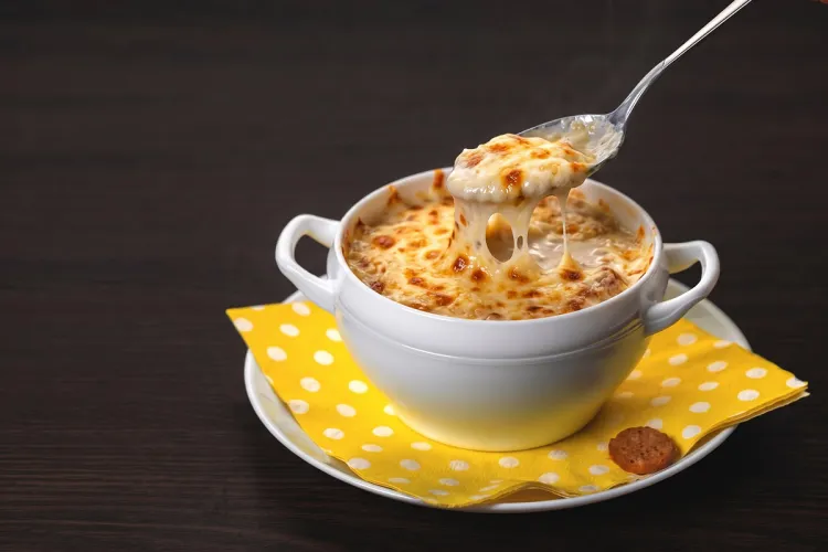 Cuenco blanco de crema de cebolla gratinada con queso fundido, con una cuchara levantando un hilo de queso, sobre una servilleta amarilla de lunares