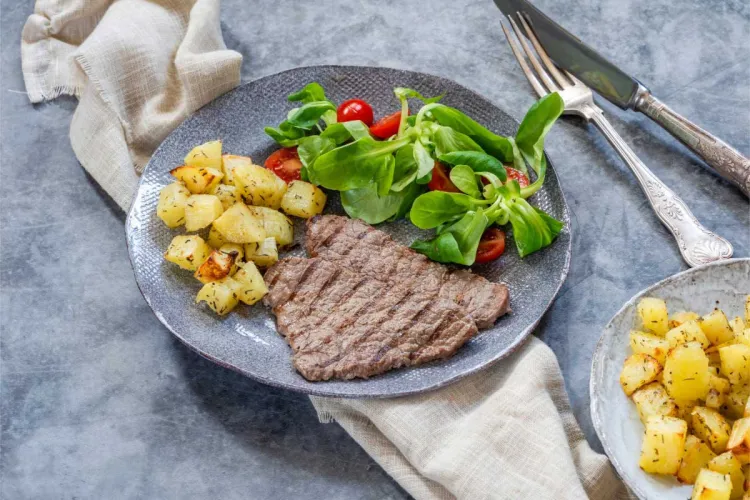 Filete a la plancha con ensalada de canónigos y tomate cherry y patatas salteadas, en un plato gris sobre una mesa de mármol