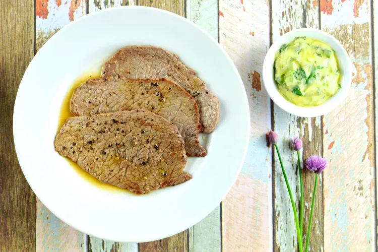 Filetes finos de vacuno a la plancha en su jugo, con un cuenco de mantequilla de albahaca al lado, vistos desde arriba sobre una mesa de madera