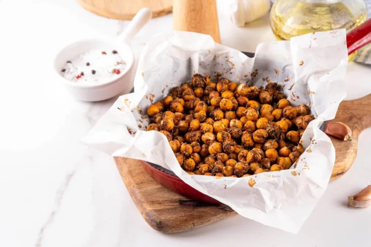 Garbanzos tostados y especiados sobre papel de horno dentro de una fuente roja, con ajos, aceite de oliva y un cuenco de sal y pimienta al fondo.