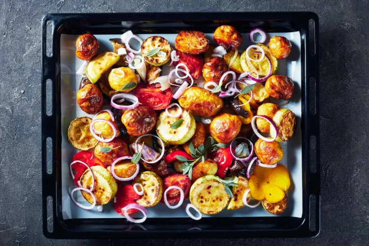 Bandeja de horno negra llena de hortalizas asadas: calabacín, tomate, patata, berenjena y pimiento, con aros de cebolla morada y hojas de salvia
