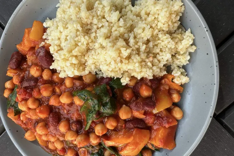 Plato hondo gris con un guiso de alubias y garbanzos en salsa de tomate al pimentón, con pimiento, judías rojas y espinacas, acompañado de un montón de bulgur, visto desde arriba sobre una mesa de madera oscura.