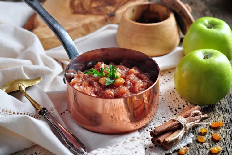 Cazo de cobre con chutney de manzana casero, decorado con una ramita de perejil y unas pasas, junto a manzanas verdes, ramas de canela y pasas sobre una mesa de madera rústica.