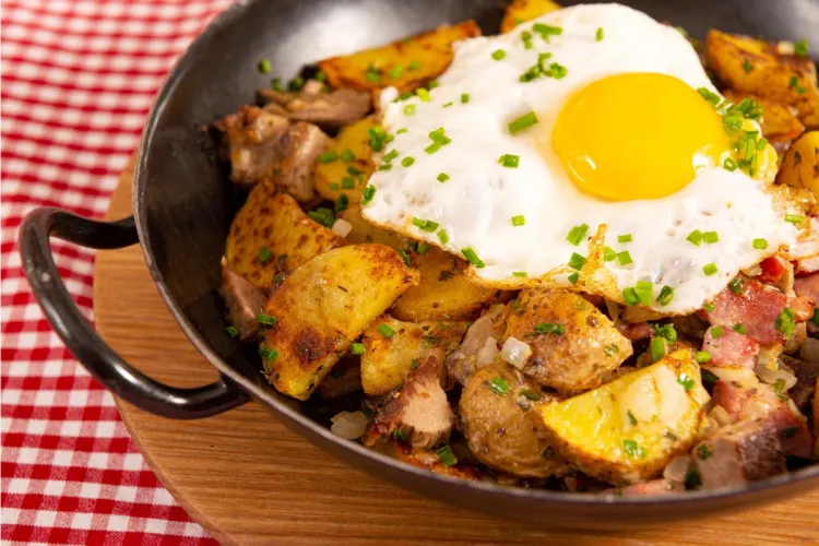 Gröstl tirolés en una sartén de hierro: patatas en rodajas doradas con tiras de carne asada y cebolla, coronado con un huevo frito de yema líquida y cebollino picado, sobre un mantel de cuadros rojos.