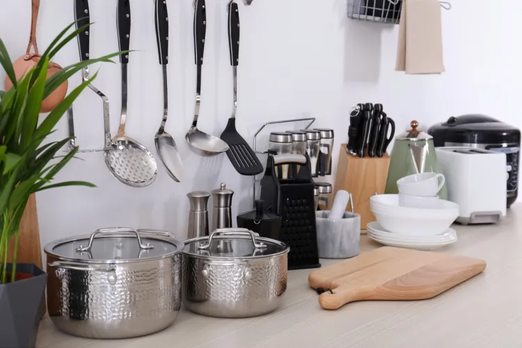 Encimera de cocina con utensilios de acero inoxidable colgados, dos ollas, tabla de madera, vajilla blanca y pequeños electrodomésticos