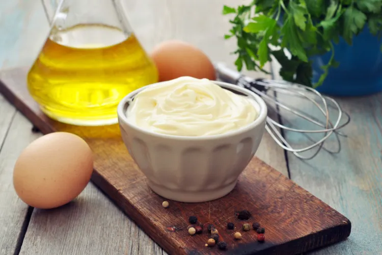 Cuenco blanco de mayonesa sobre una tabla de madera, con huevos, aceite en una jarra, unas varillas y granos de pimienta