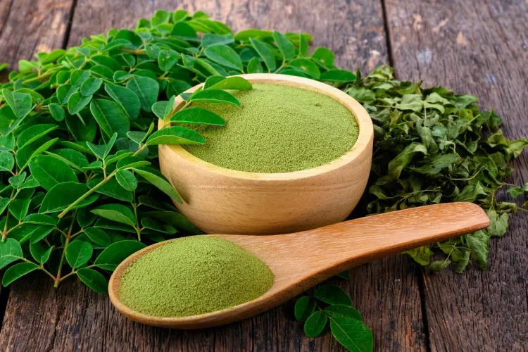Cuenco y cuchara de madera con polvo de moringa verde, rodeados de hojas frescas y secas de moringa sobre una mesa de madera rústica.