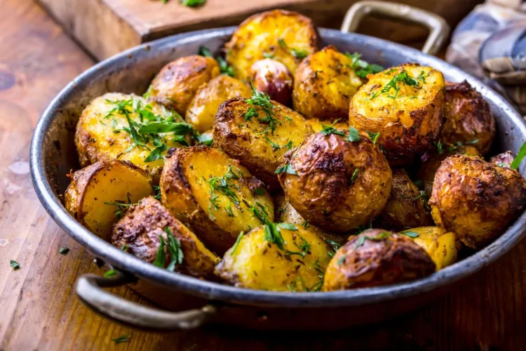 Mitades de patata nueva asadas y doradas, con la piel puesta y espolvoreadas con perejil y eneldo picados, en una sartén metálica sobre una mesa de madera