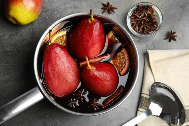 Vista cenital de un cazo con peras al vino tinto, enteras y teñidas de rojo, cocidas en vino con rodajas de naranja, ramas de canela, anís estrellado y clavo; al lado, un cucharón, una pera fresca y un cuenco de anís estrellado.
