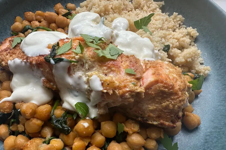 Lomo de salmón especiado y dorado, napado con salsa de yogur y espolvoreado de perejil, sobre garbanzos salteados con espinacas y con cuscús al lado, en un plato azul